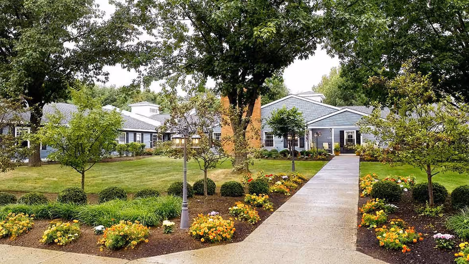 A paved walkway leading through a landscaped garden with colorful flowers and trimmed bushes, flanked by trees, towards a single-story blue building with white trim and multiple windows under a cloudy sky.