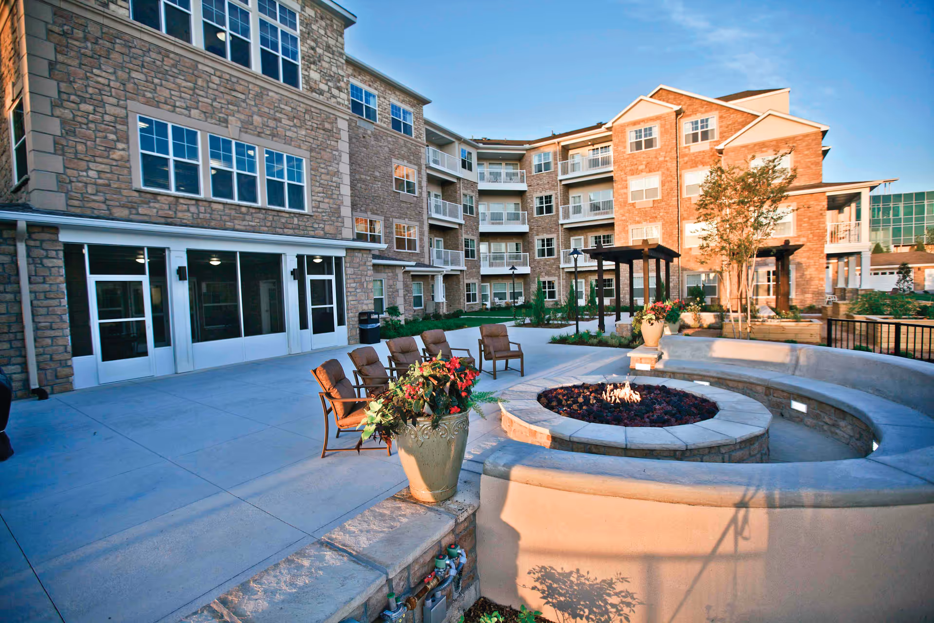 Outdoor patio area of a senior living facility with a circular stone fire pit surrounded by curved concrete seating and several chairs. The building in the background is multi-story with stone and brick exterior walls, multiple windows, and balconies. There are potted plants and some landscaping around the patio area under a clear blue sky.