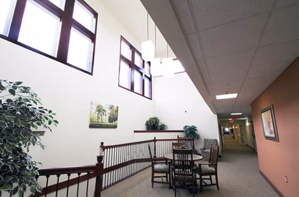 Interior hallway of Kennybrook Village with a seating area featuring a round table and four chairs. The space is well-lit with large windows near the ceiling, hanging pendant lights, and decorated with plants and framed artwork on the walls.
