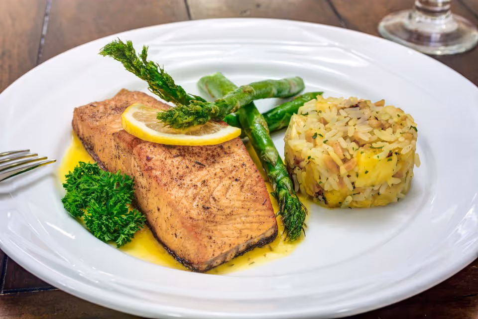 A plated meal featuring a cooked salmon fillet topped with a lemon slice and garnished with parsley, served with asparagus spears and a molded serving of rice with herbs and small pieces of vegetables on a white plate.