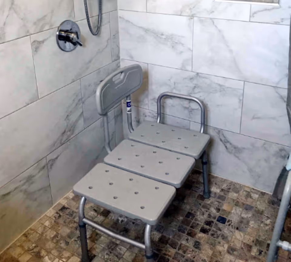 Shower area with a gray three-panel shower bench and backrest against marble-look tiled walls and a handheld showerhead.
