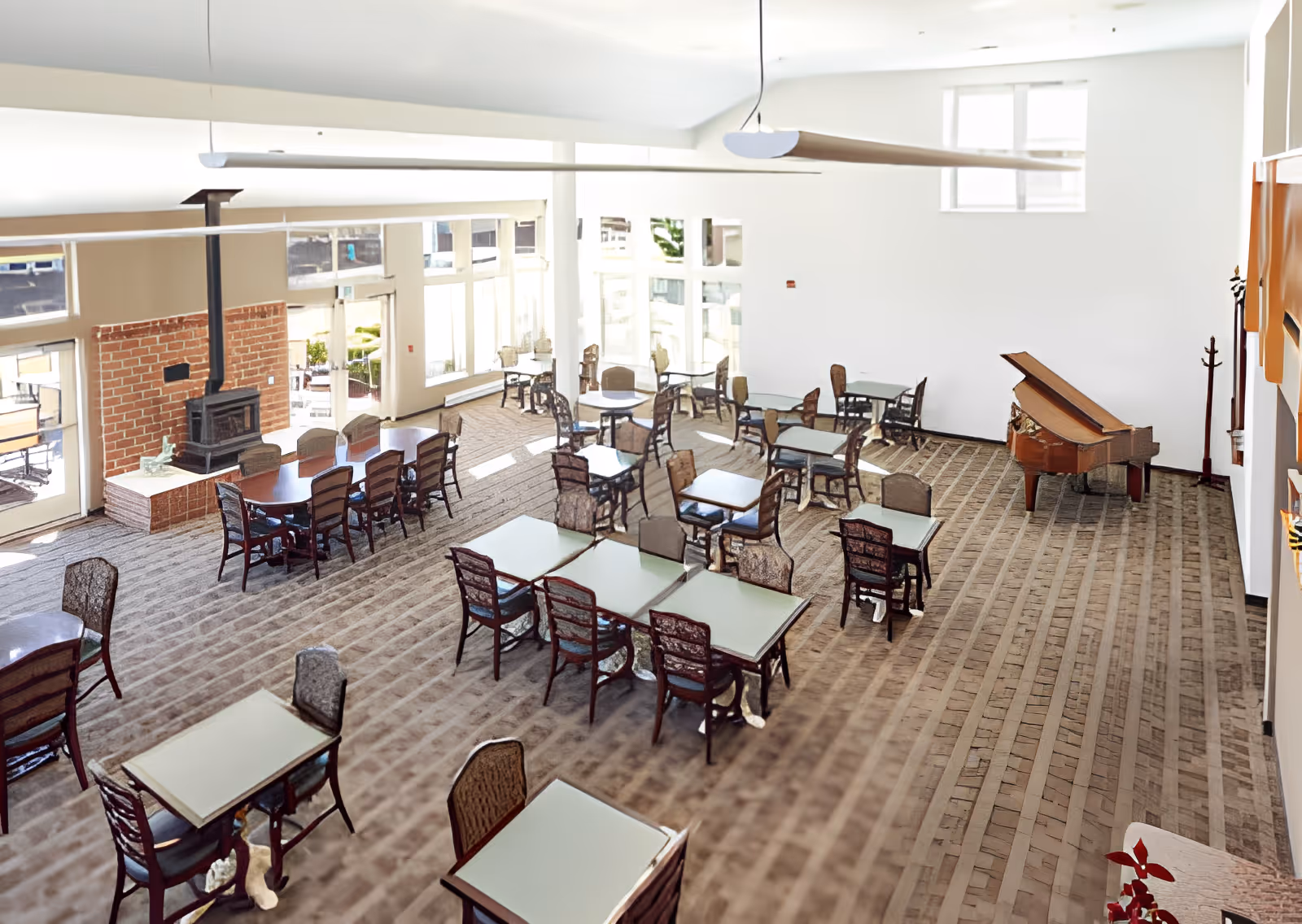A spacious dining room with multiple tables and chairs arranged neatly on a patterned carpet. There is a brick fireplace on the left side and a grand piano on the right side near the wall. Large windows allow natural light to fill the room.