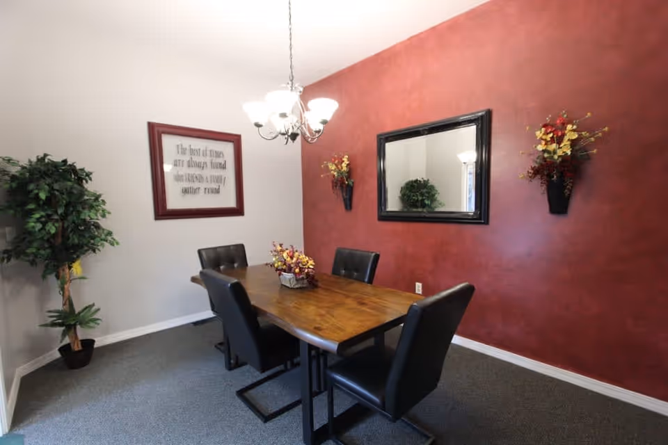 A dining room with a wooden table surrounded by four black cushioned chairs. The room has a red accent wall with a large black-framed mirror and two wall-mounted flower arrangements. A chandelier with five lights hangs above the table. On the opposite wall, there is a framed quote and a tall potted plant in the corner.