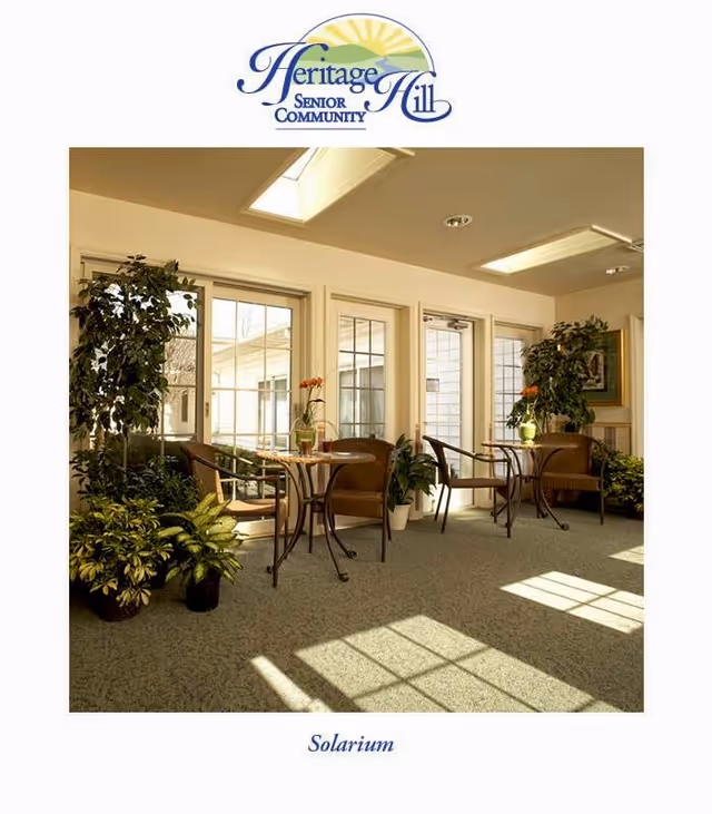 A bright solarium room with large windows and glass doors allowing natural light to fill the space. The room features small round tables with wicker chairs, potted plants, and a framed picture on the wall, creating a cozy and inviting atmosphere.