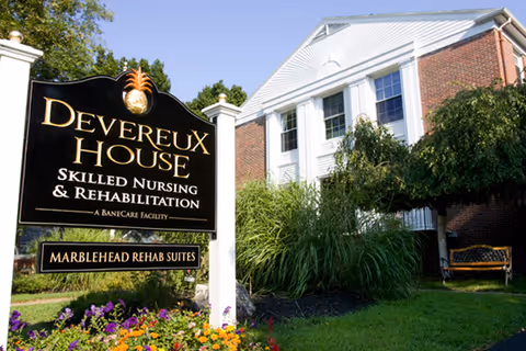 Outdoor view of Devereux House Skilled Nursing & Rehabilitation facility sign with flowers and greenery in front, and a brick building with white columns and windows in the background.