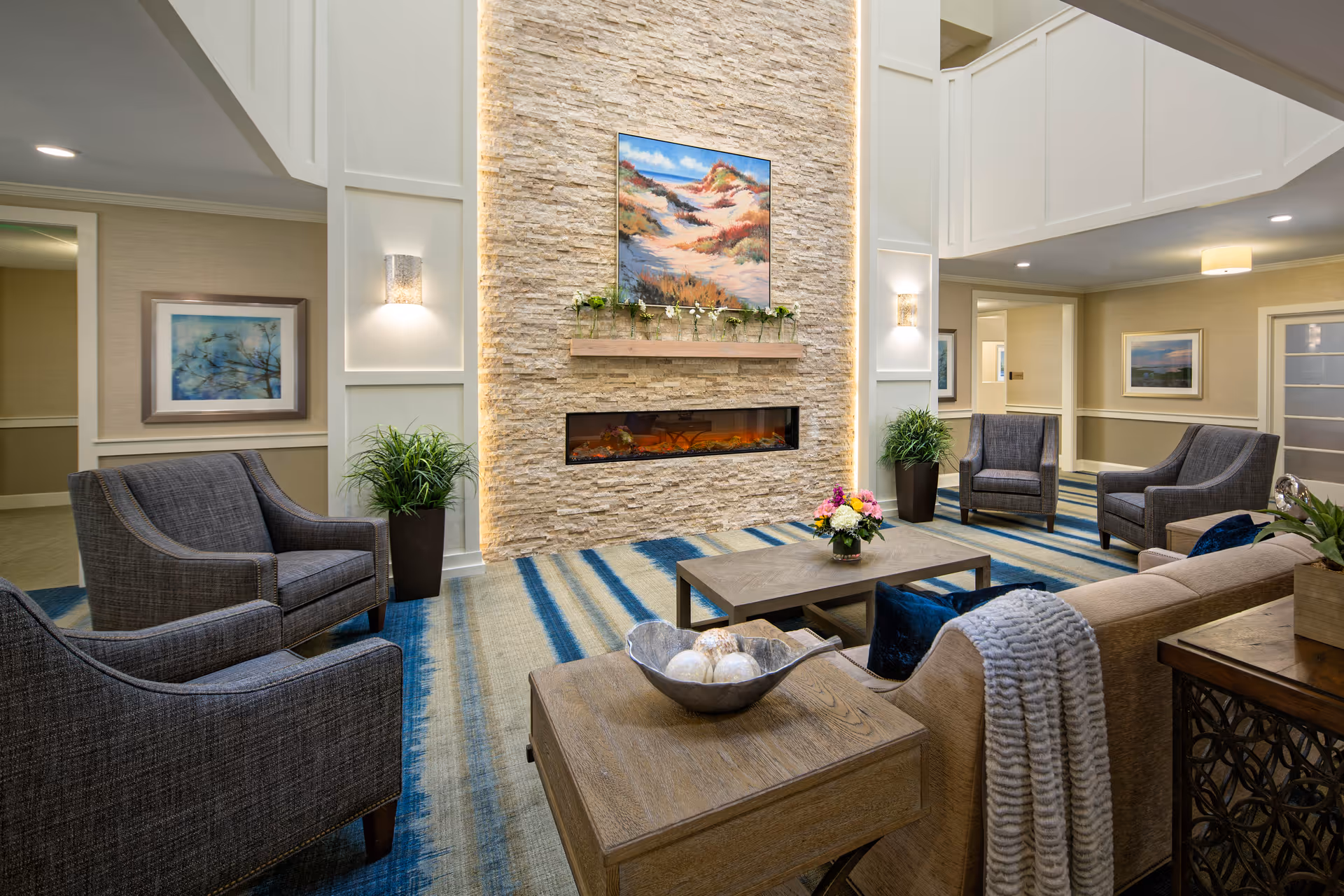 A spacious, well-lit lounge with armchairs and sofas arranged around a modern stone fireplace and coffee table.