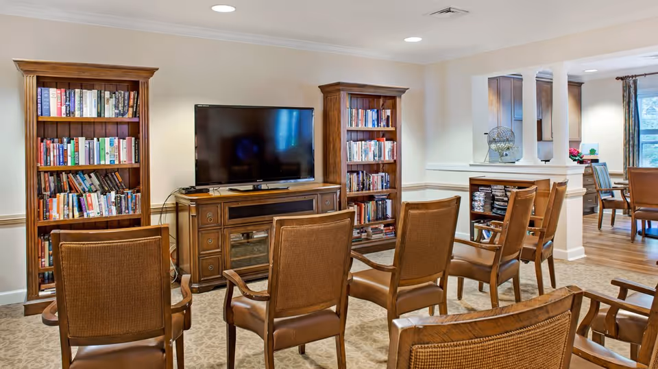 A cozy common area with several wooden chairs arranged facing a large flat-screen TV on a wooden cabinet. Two tall wooden bookshelves filled with books flank the TV. The room has beige walls, carpeted flooring, and recessed ceiling lights. In the background, there is a partial view of a dining area with wooden chairs and a window with curtains.