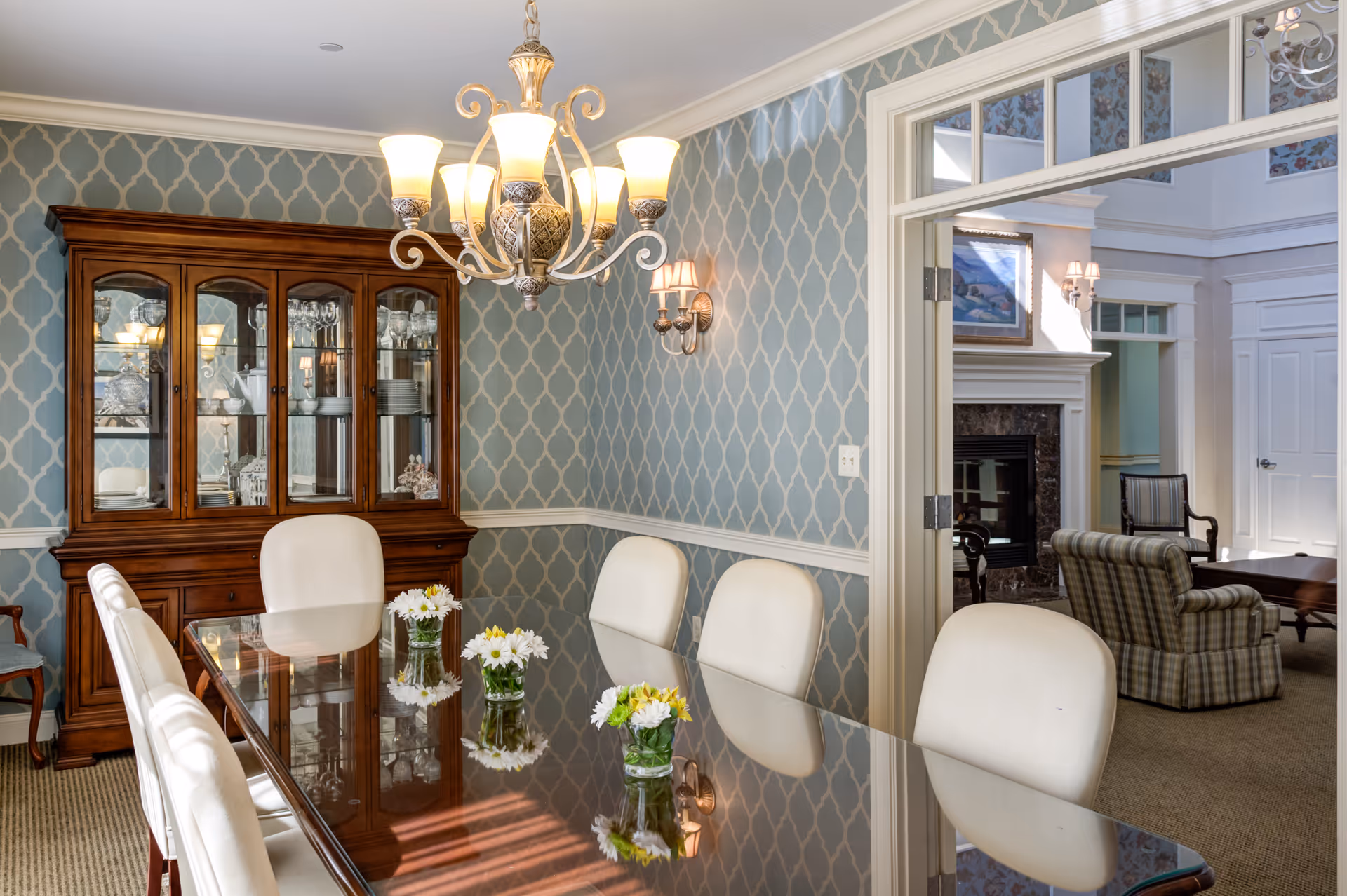 A formal dining room with a long glass-top table surrounded by white upholstered chairs. On the table are small vases with white and yellow flowers. The walls have a blue patterned wallpaper with white trim. A wooden china cabinet with glass doors displays dishes and glassware. A chandelier with multiple lights hangs above the table. Through an open doorway, a living room area with armchairs and a fireplace is visible.