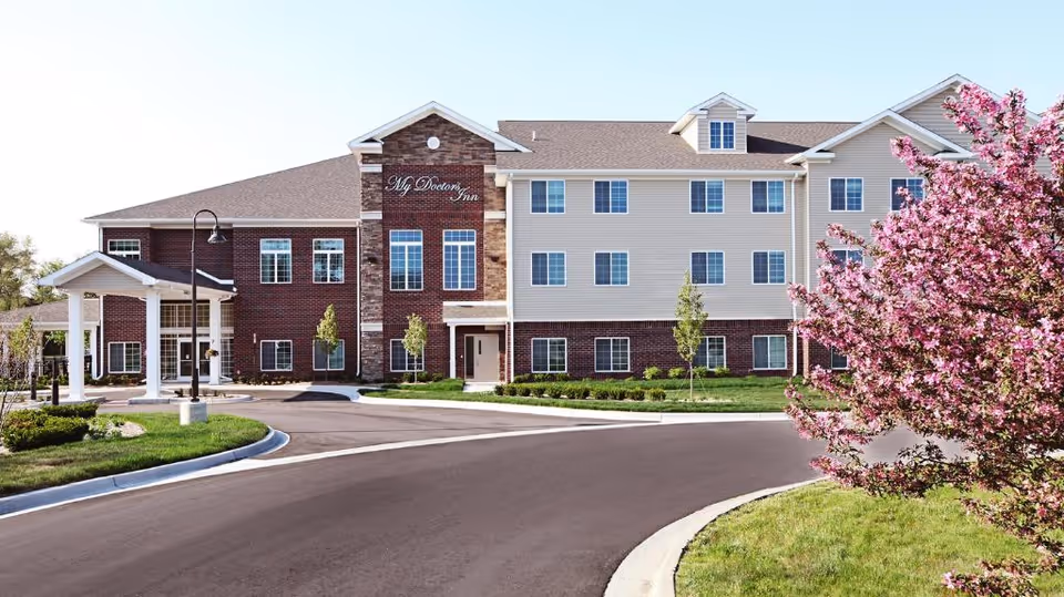 Three-story assisted living building facade with a circular driveway and a pink blossoming tree near the entrance.