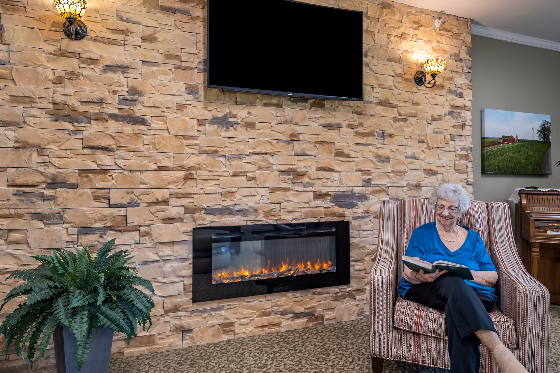 An elderly woman with white hair and glasses sits comfortably in a striped armchair, reading a book. She is in a cozy room with a stone wall featuring a modern electric fireplace and a mounted flat-screen TV above it. A green potted plant is on the left, and a wooden piano with a landscape painting above it is visible in the background.