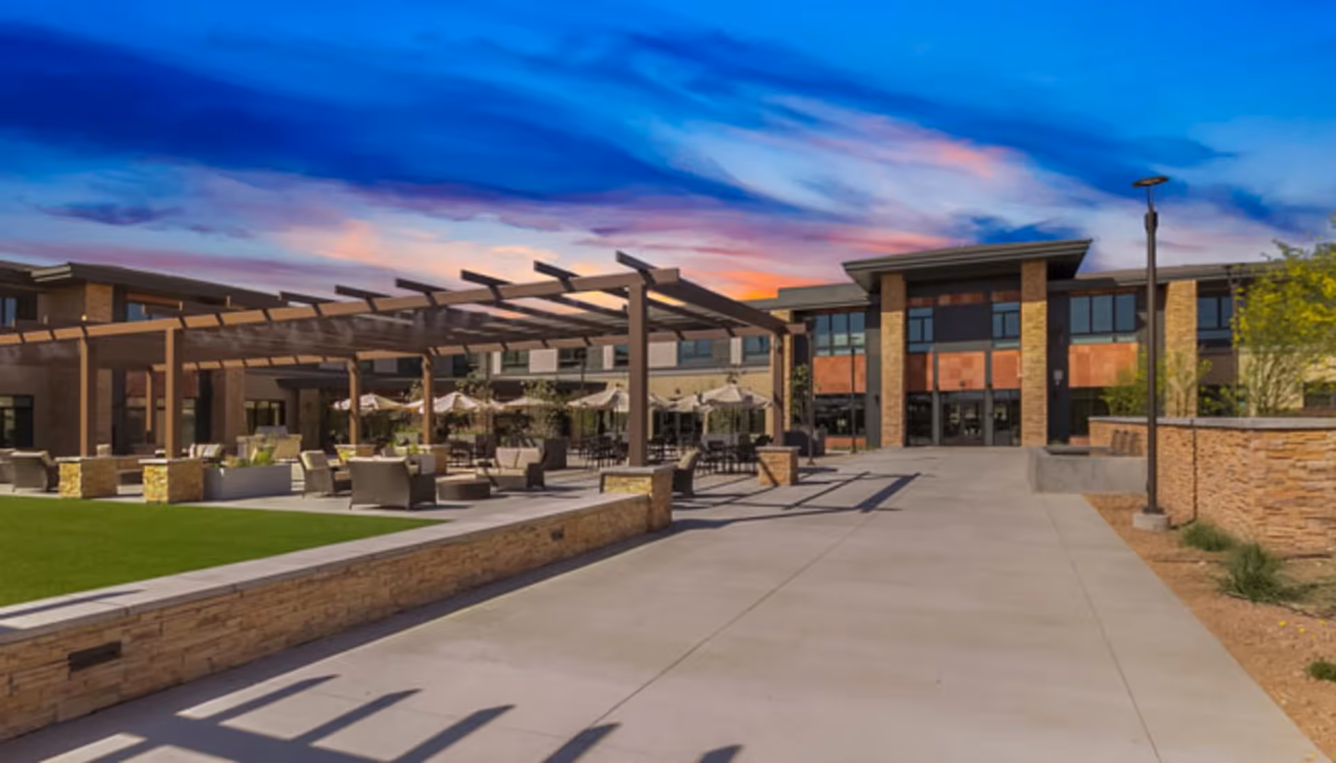 Outdoor patio area of a senior living facility with seating arrangements under pergolas and umbrellas, adjacent to a modern building under a colorful sunset sky.