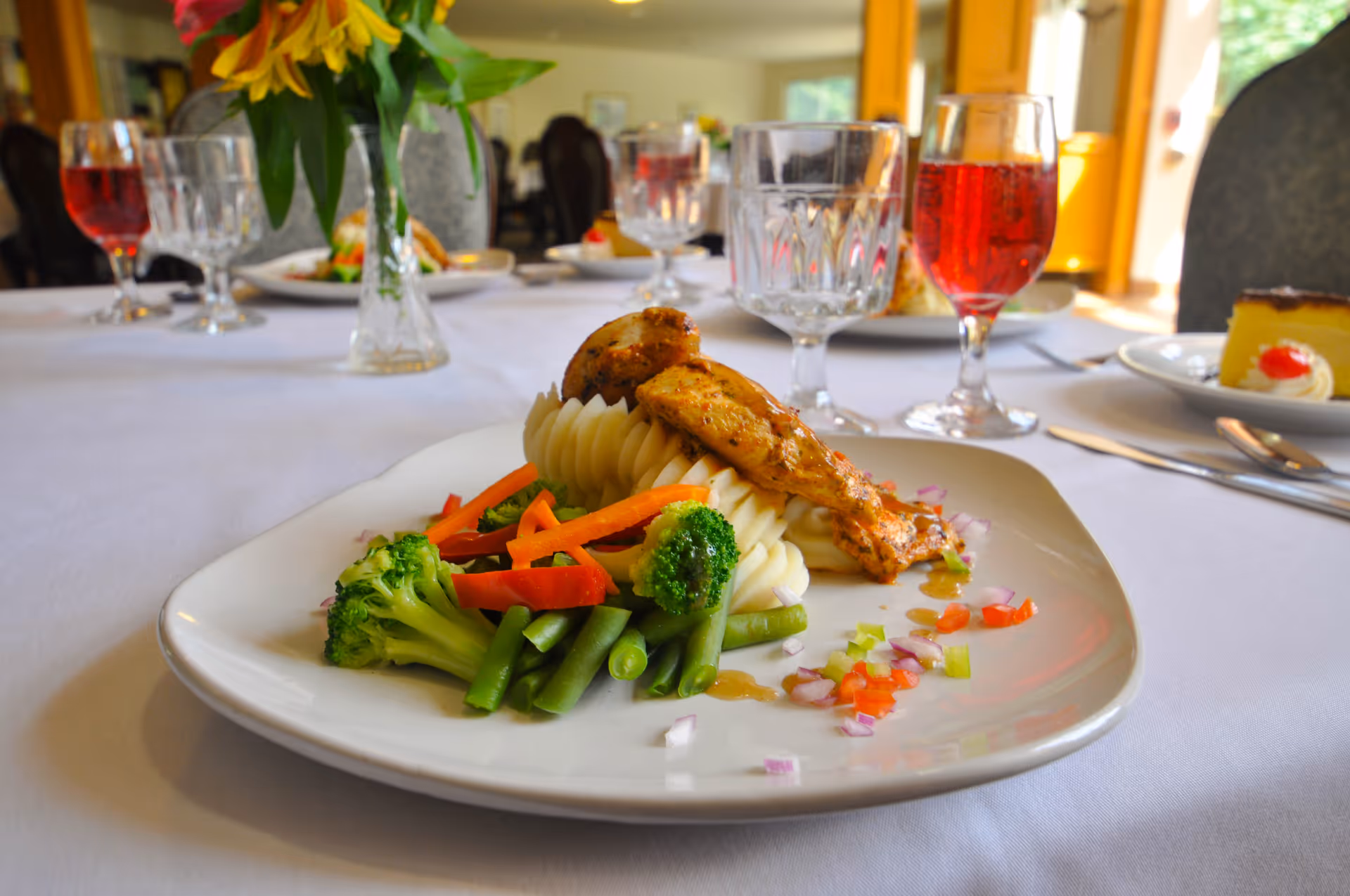A plated meal featuring grilled chicken breast served on a bed of mashed potatoes with steamed vegetables including broccoli, green beans, and carrots, garnished with finely chopped red and green peppers. The plate is on a white tablecloth with a vase of yellow flowers, crystal glasses, and a glass of red beverage in the background, set in a dining room.