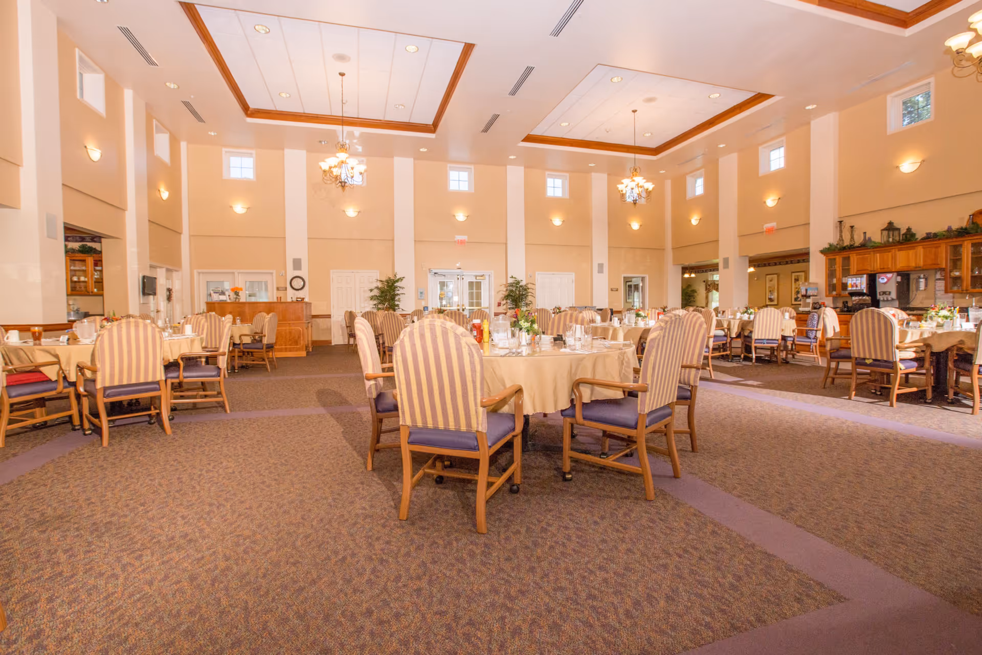 Bright, spacious dining room with round tables set for meals, striped upholstered chairs, chandeliers, and high ceilings.