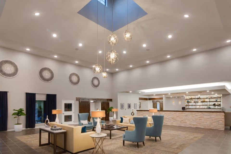 A spacious and modern living room area with high ceilings and a skylight. The room features beige sofas, blue armchairs, several table lamps, and decorative wall plates. There is a reception or bar counter with shelves and pendant lights in the background.