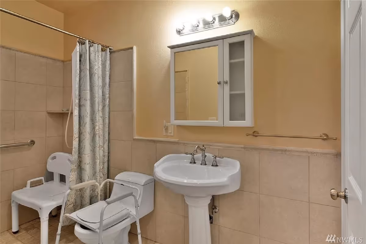 Bathroom with beige tiled walls and floor, featuring a white pedestal sink with a mirror cabinet above it, a toilet with a white shower chair placed over it, another white shower chair inside a tiled shower area with a floral shower curtain, and a towel bar on the wall.