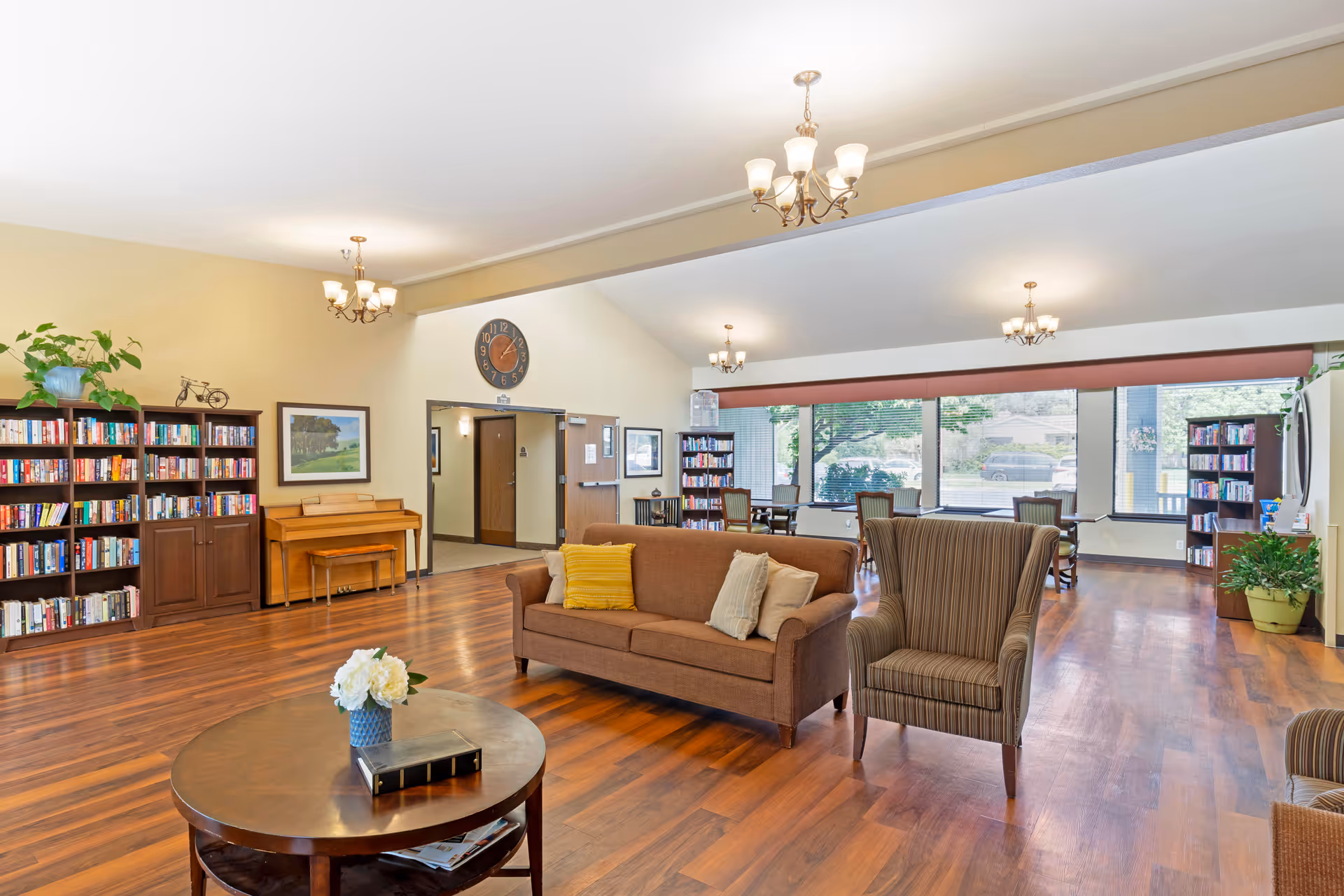 A spacious and well-lit common area with wooden flooring, featuring a brown sofa with cushions, a striped armchair, a round wooden coffee table with a flower vase and a book, bookshelves filled with books, a piano, and several tables and chairs near large windows that let in natural light.
