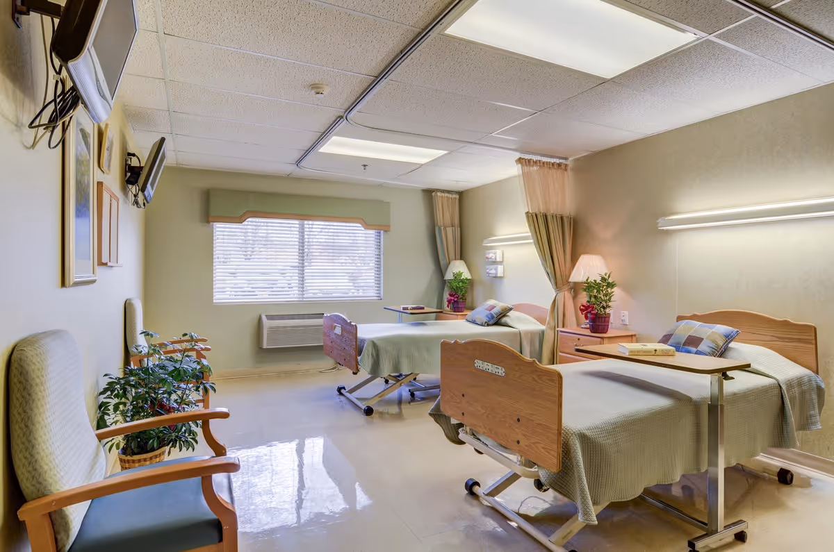 A bright shared patient room with two hospital-style beds, bedside tables, chairs, lamps, and plants.