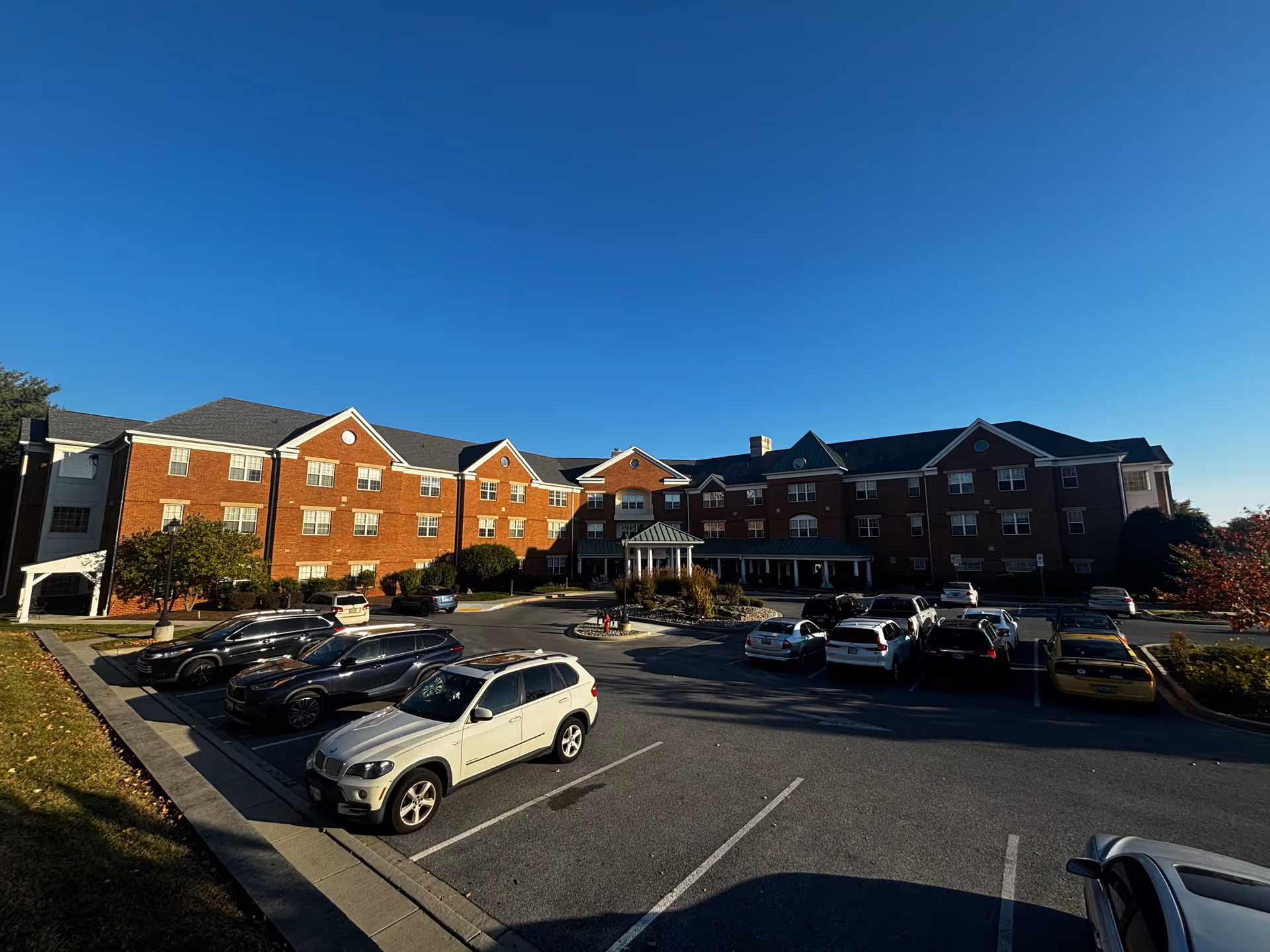 Exterior view of a large three-story brick building with multiple windows and a covered entrance. Several cars are parked in the parking lot in front of the building under a clear blue sky.