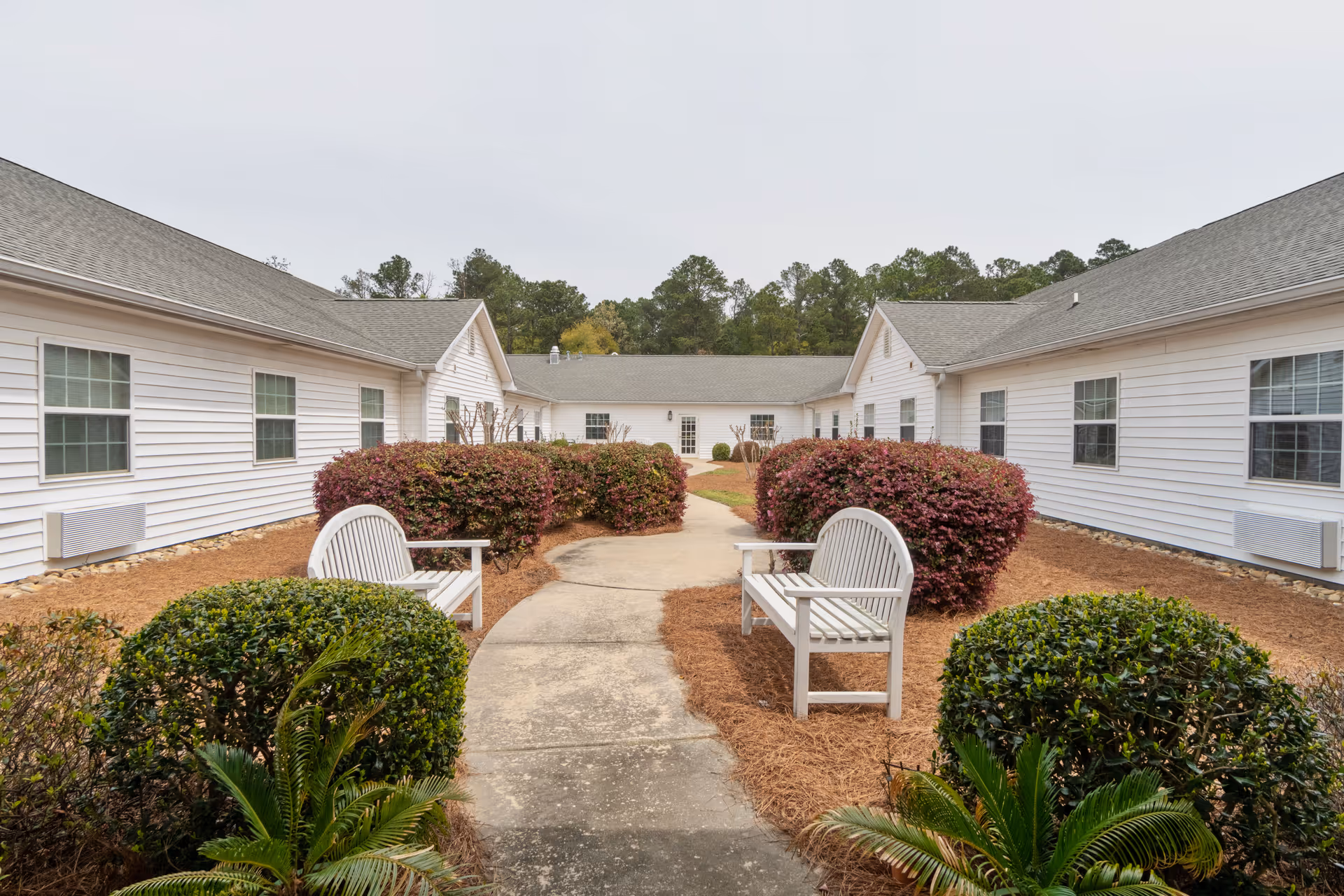 Outdoor courtyard area at Merryvale Assisted Living featuring a paved walkway flanked by white benches and neatly trimmed bushes, surrounded by single-story white buildings with multiple windows under a cloudy sky.