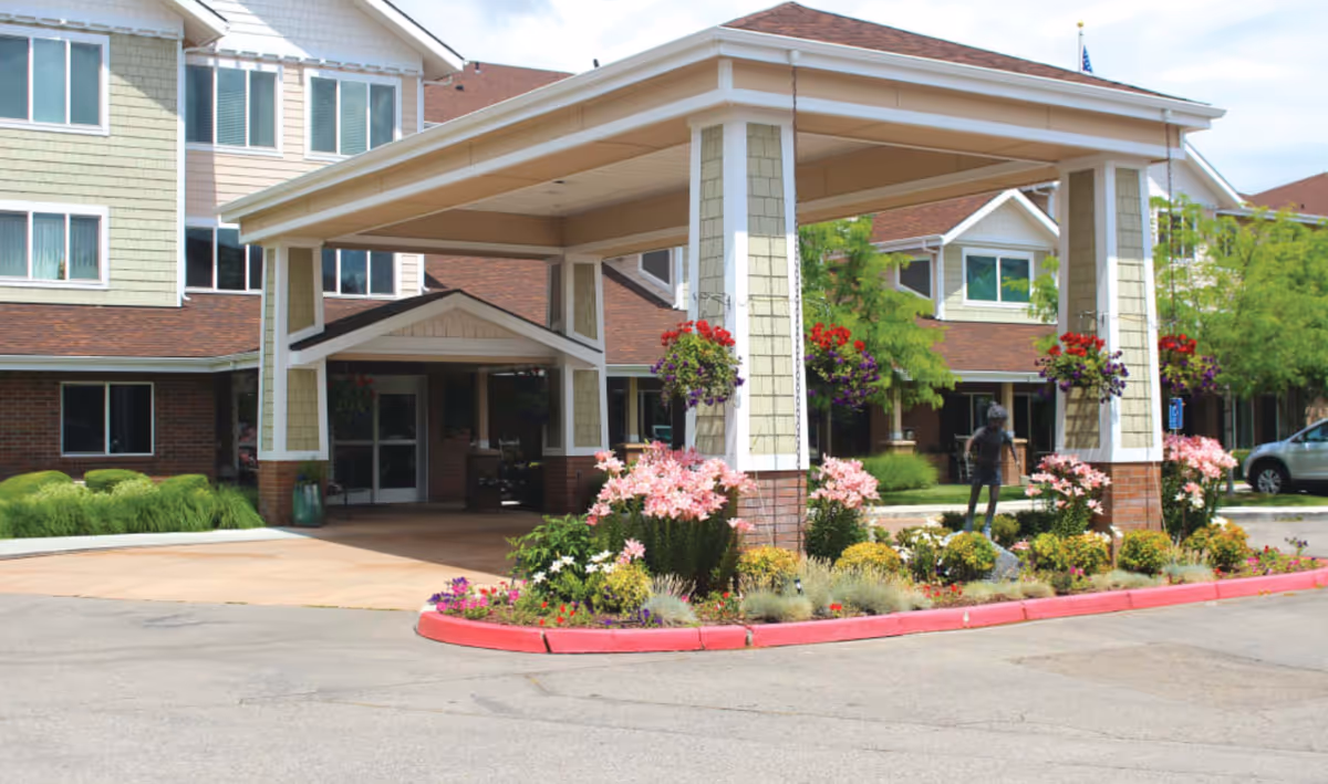 Front entrance of a senior living facility with a covered porte-cochere, landscaped flowerbeds and hanging baskets.