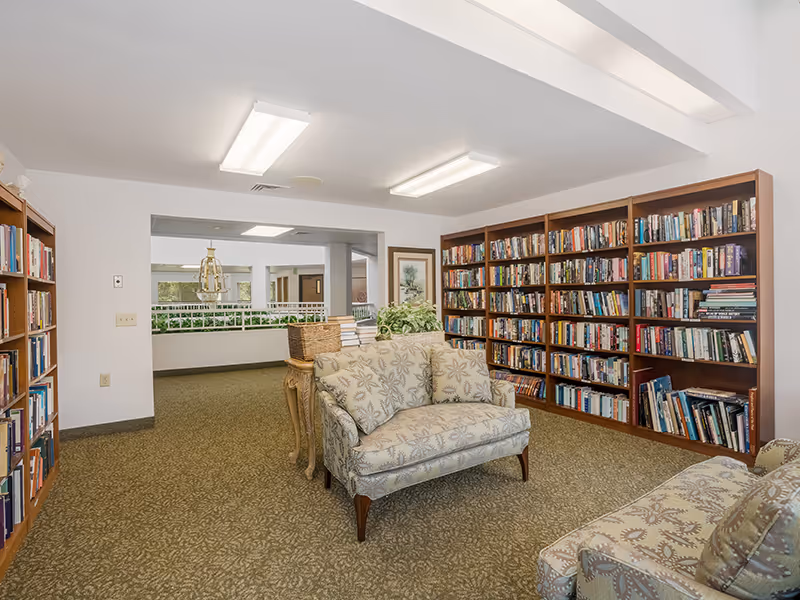 A cozy reading area in a senior living facility with two patterned upholstered loveseats facing each other. Surrounding the seating are tall wooden bookshelves filled with books. The room has a carpeted floor and bright overhead lighting. In the background, there is an open space with a railing and some greenery.