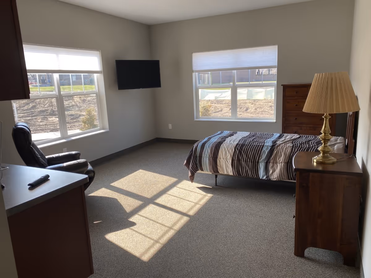 A bright bedroom with two large windows letting in natural light. The room contains a single bed with a striped comforter, a wooden dresser with a brass lamp on top, a wall-mounted flat-screen TV, a black leather recliner chair, and a countertop with a remote control on it. The walls are painted light gray and the floor is carpeted.