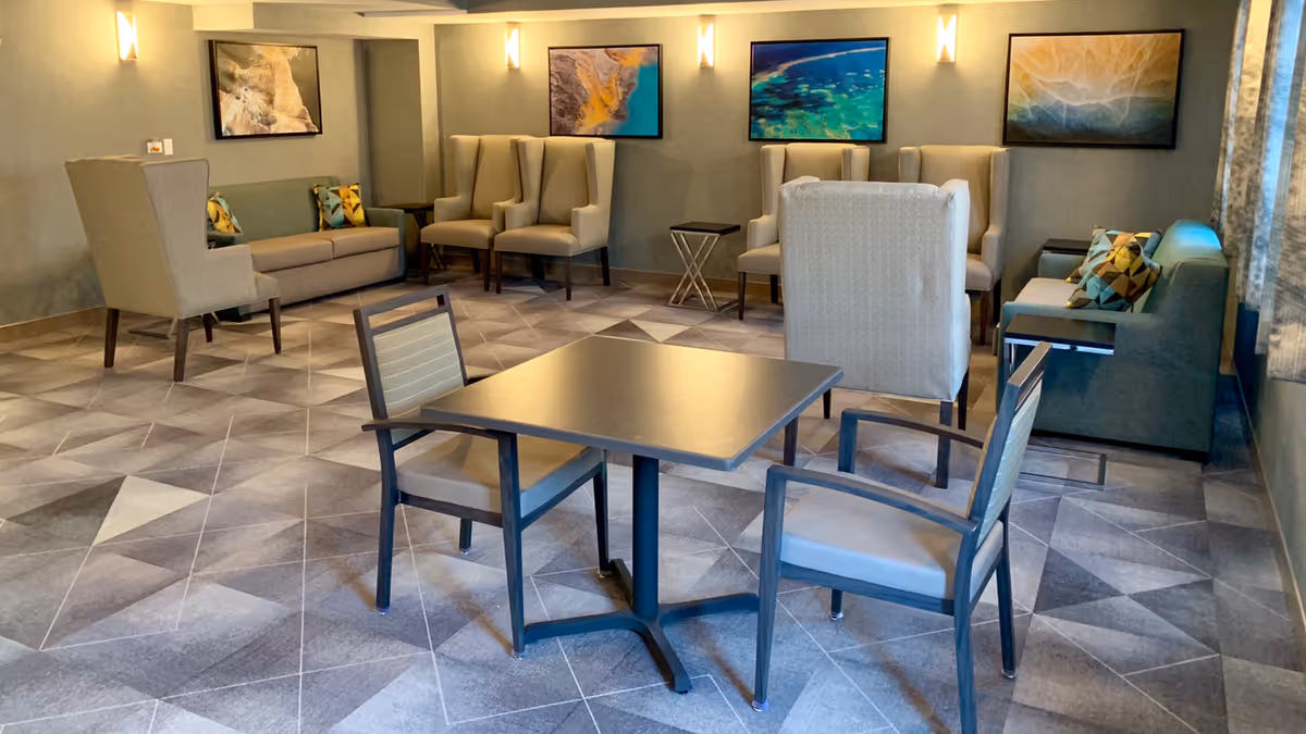 A well-lit supportive living facility common area with a square table and two chairs in the foreground. The room features multiple seating options including sofas, armchairs, and high-back chairs arranged along the walls. The walls are decorated with framed abstract art, and soft wall sconces provide warm lighting. The floor has a geometric patterned carpet.