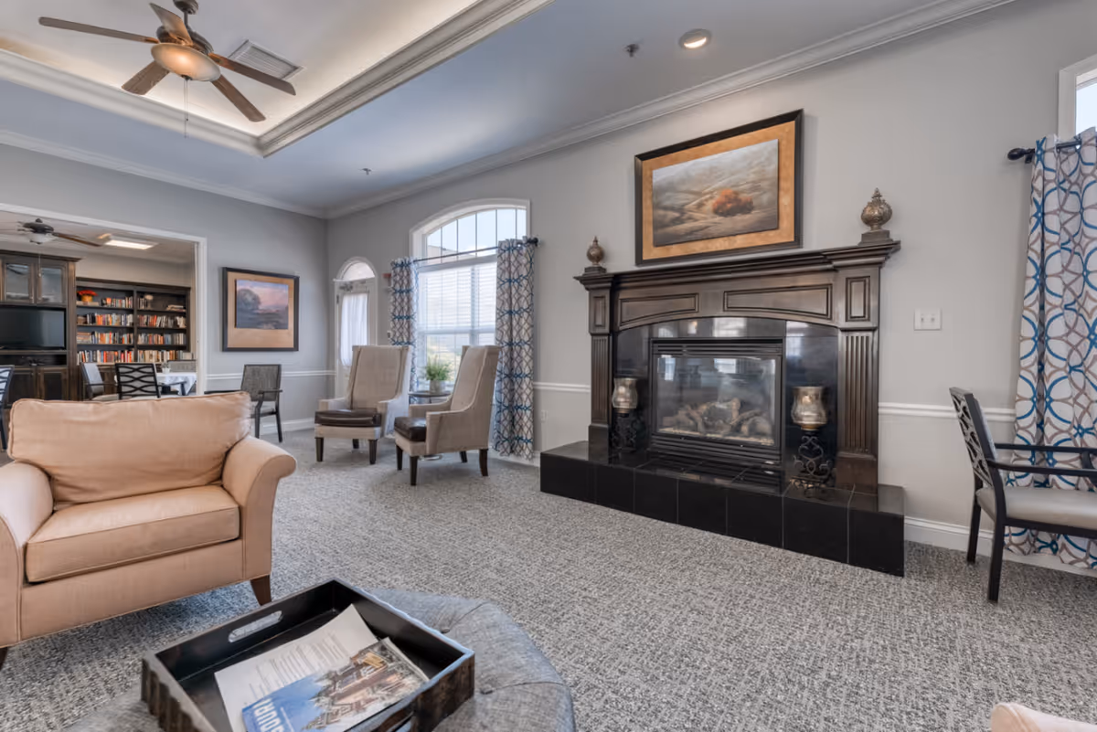 A cozy living room area with a beige armchair, two gray upholstered chairs near a window with patterned curtains, and a dark wooden fireplace with a painting above it. The room has a gray carpet, light gray walls, and a ceiling fan. In the background, there is a bookshelf and a dining area with chairs and a table.