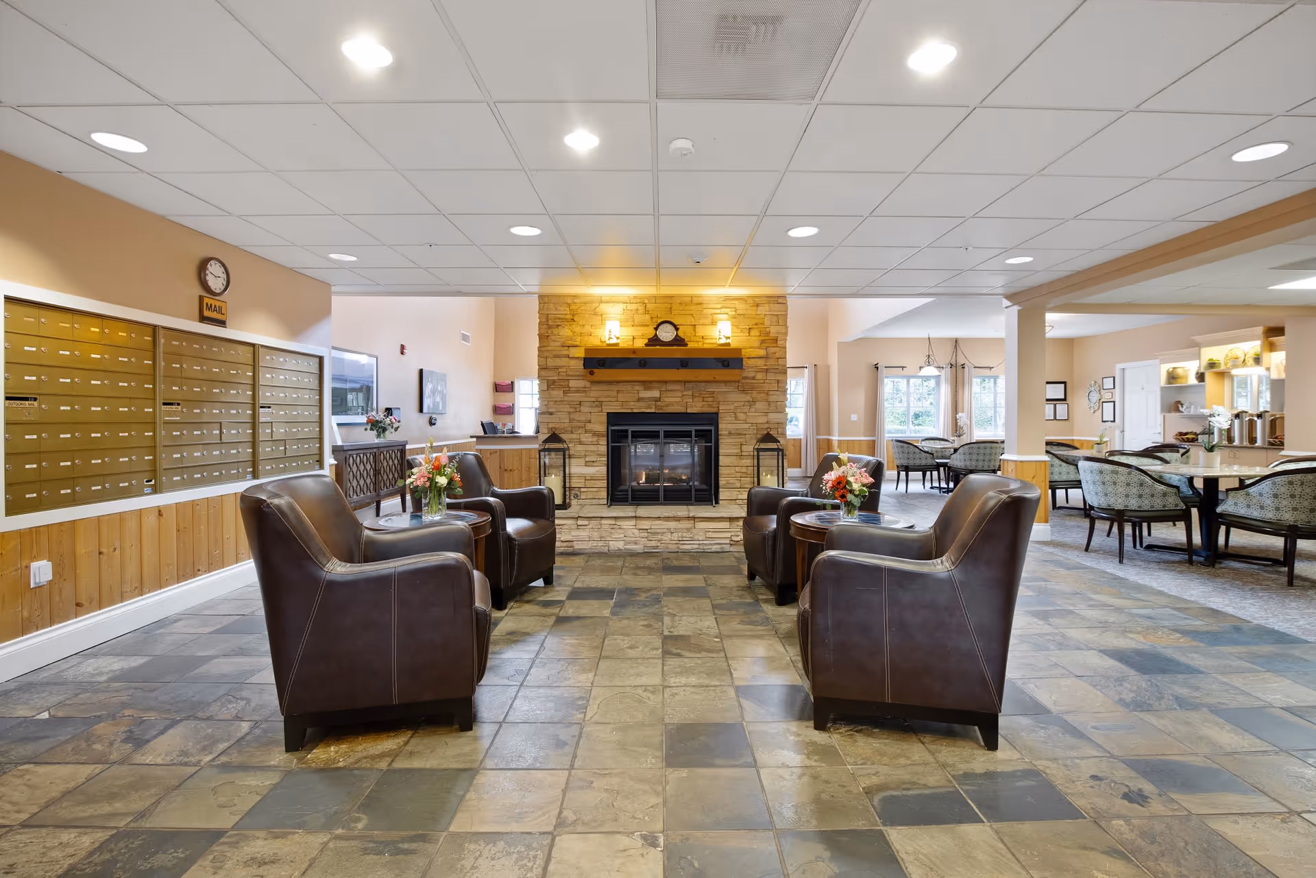 A spacious senior living facility common area with four brown leather armchairs arranged around two small round tables with flower vases. A stone fireplace with a clock and two lanterns is centered on the far wall. To the left, there is a wall of mailboxes labeled 'MAIL' with a clock above. To the right, there are dining tables and chairs near large windows letting in natural light. The floor is tiled with a mix of earth tones.