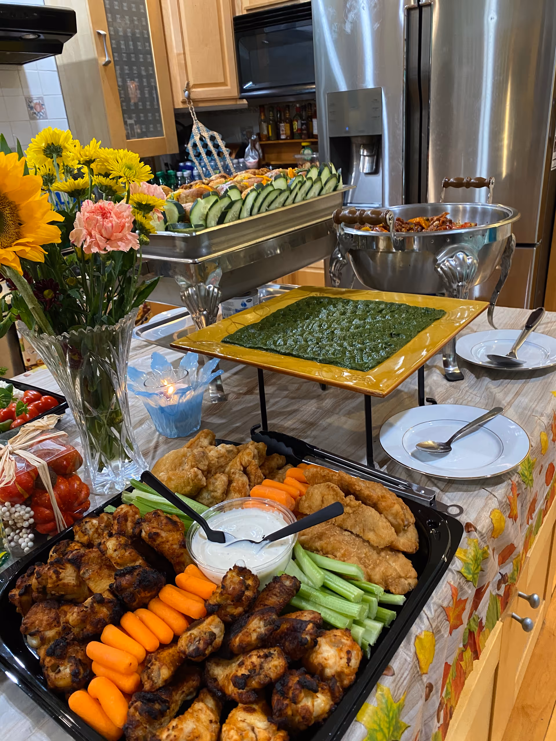 A kitchen counter set up with a variety of food trays including grilled chicken wings, baby carrots, celery sticks, fried chicken strips, a bowl of white dipping sauce, a tray of sliced cucumbers, and a green dip on a yellow square plate. There is a vase with sunflowers and pink carnations, a lit candle in a blue holder, and plates with spoons on the counter. The background shows wooden kitchen cabinets and a stainless steel refrigerator.