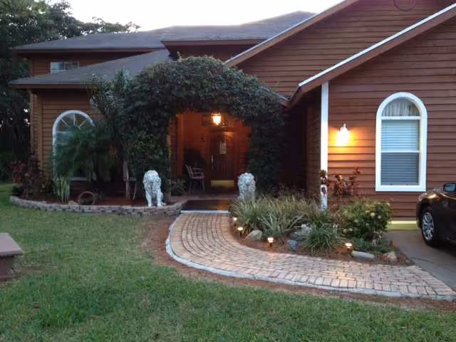 Front exterior of a brown single-story house with a curved brick walkway, an arched vine-covered entrance, and two lion statues flanking the path.