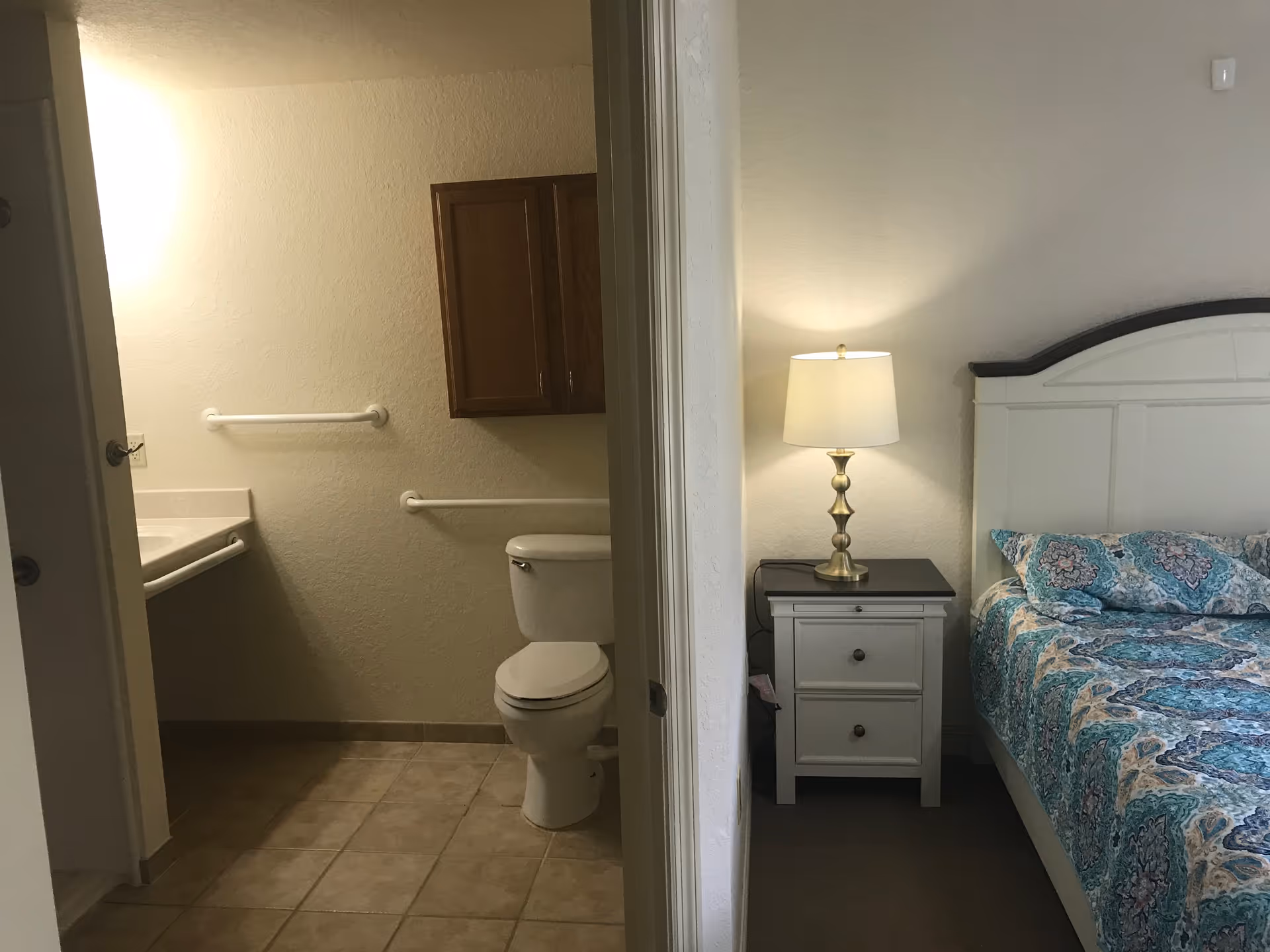 View of a bedroom and an adjacent bathroom in a senior living facility. The bedroom features a bed with a patterned blue and white bedspread, a white nightstand with two drawers, and a brass table lamp with a white shade. The bathroom has beige tiled flooring, a white toilet, a sink with a countertop, grab bars on the walls, and a wooden cabinet mounted above the toilet.