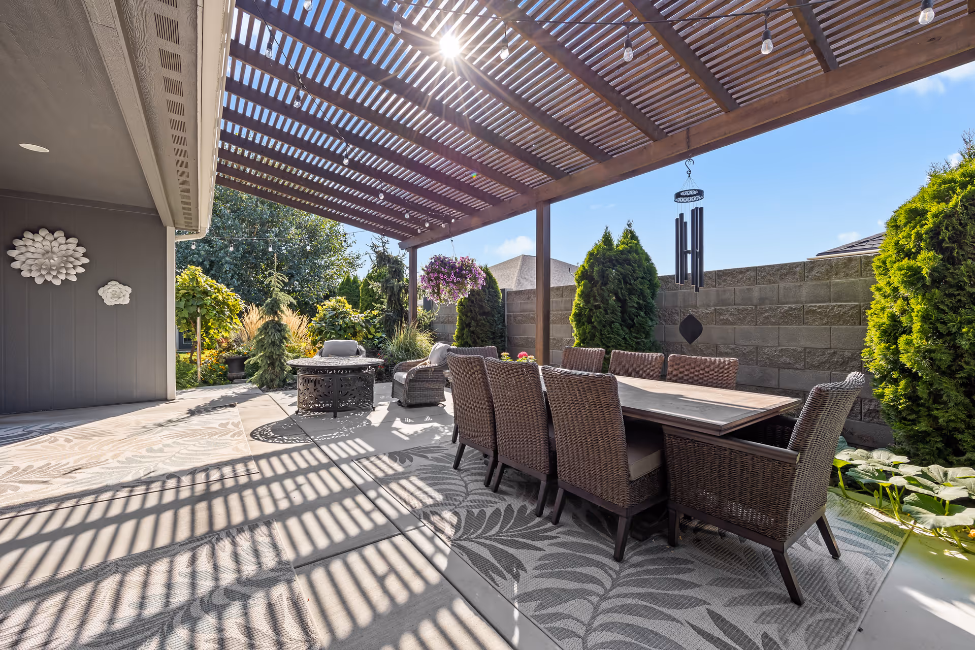 Covered outdoor patio with a long dining table and wicker chairs under a wooden pergola casting striped shadows, with additional seating and surrounding greenery.