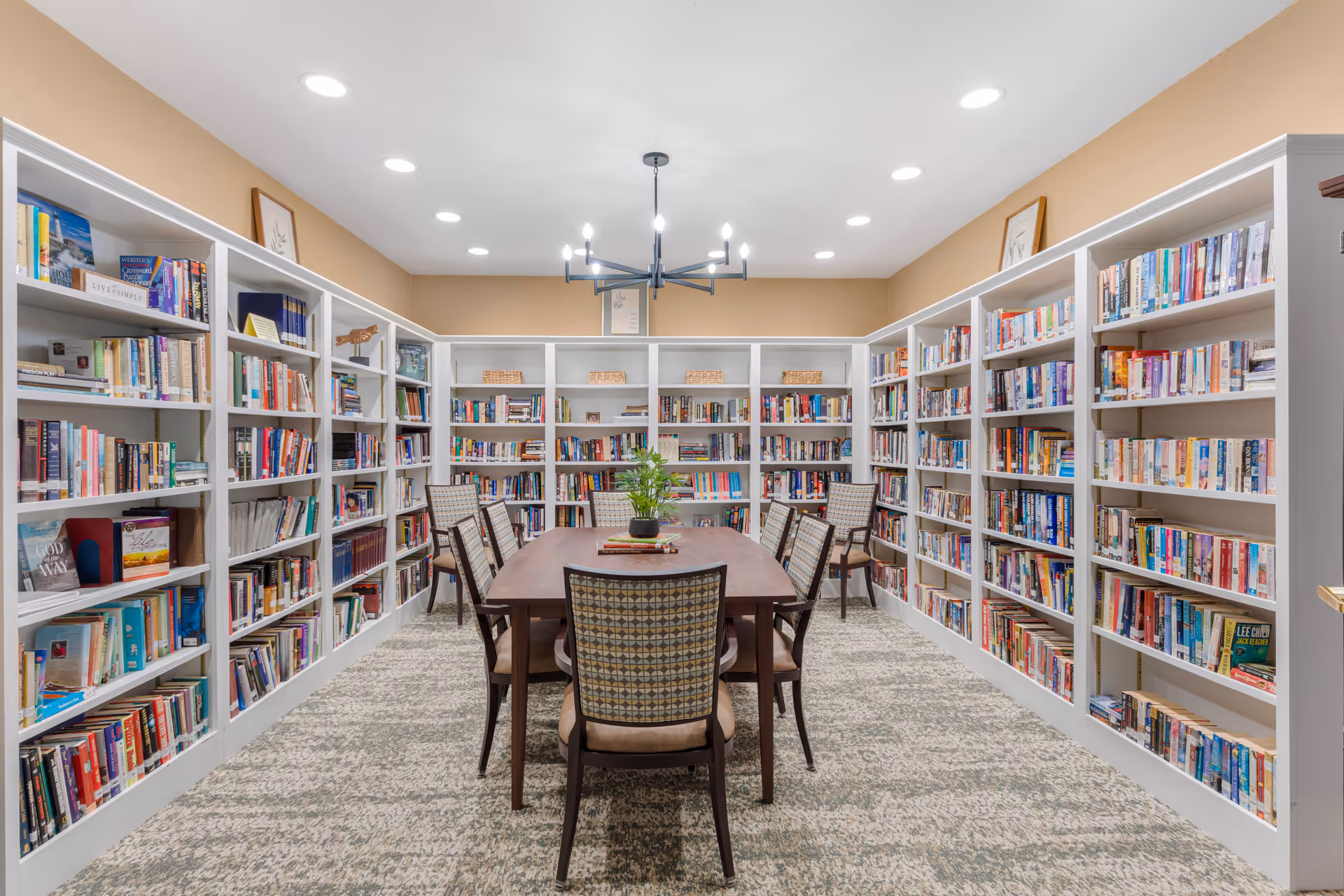 A well-lit library room with beige walls and carpeted floor. The room features white bookshelves filled with books lining three walls. In the center, there is a wooden table surrounded by eight upholstered chairs with patterned backs. A modern chandelier hangs from the ceiling above the table, and a small plant and some books are placed on the table.