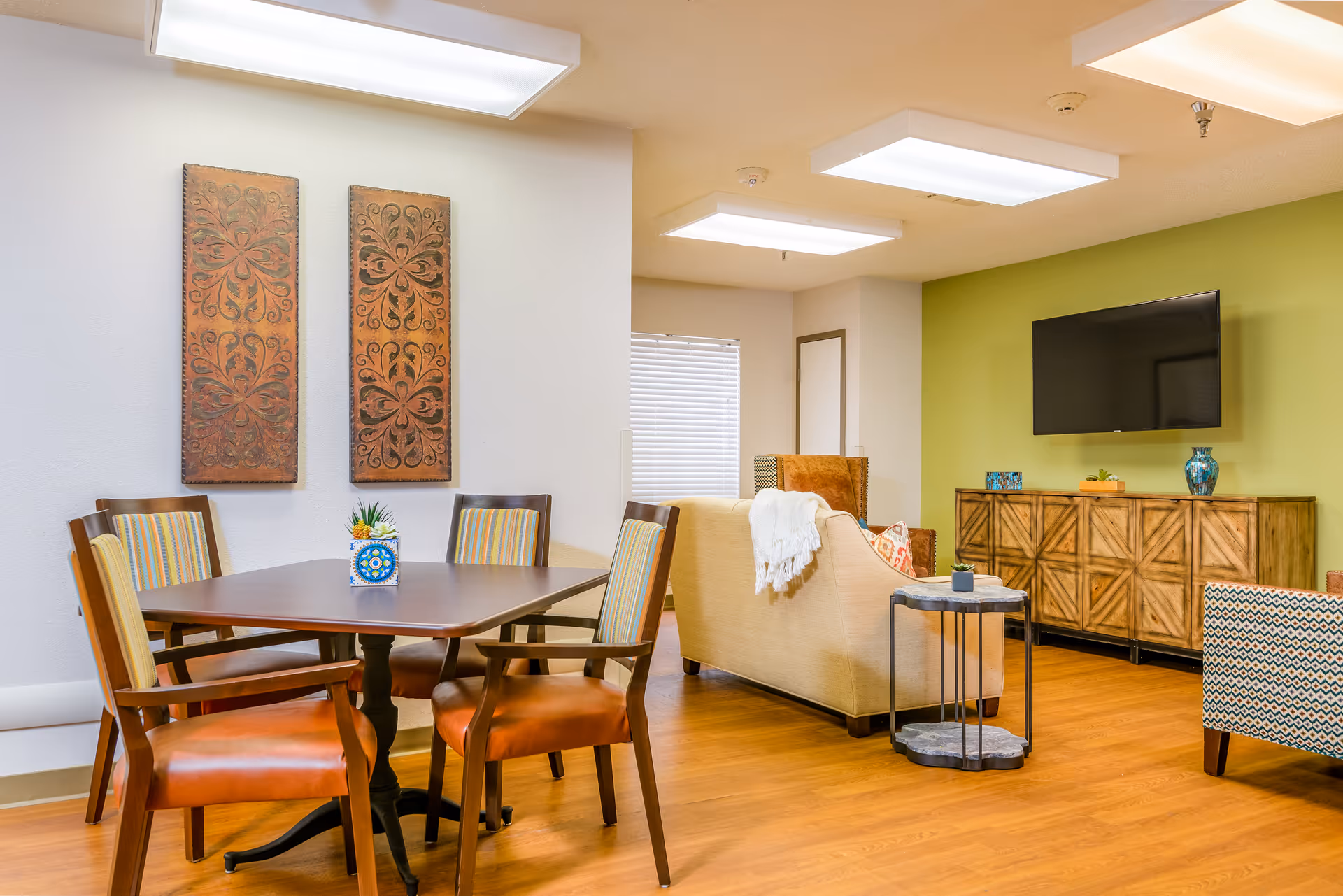 A bright and cozy common area in a senior living facility featuring a wooden dining table with four chairs, a beige couch with a white throw blanket, a patterned armchair, a wooden sideboard with decorative items, and a wall-mounted flat-screen TV. The walls are painted white and green, and the floor is hardwood.