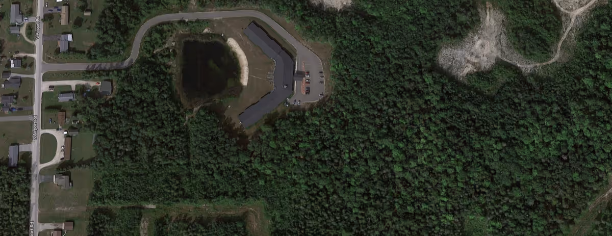 Aerial view of a curved building and parking area surrounded by dense forest with nearby houses and roads.