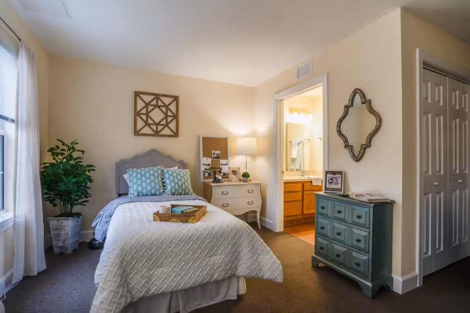 A cozy bedroom with a single bed covered in a white quilt and two blue patterned pillows. A wooden tray with a cup and a book rests on the bed. To the left, there is a large window with sheer white curtains and a potted plant. Next to the bed is a small white nightstand with a lamp, a mirror, and some decorative items. On the right side of the room, there is a green dresser with a framed photo and a decorative mirror above it. An open door reveals a bathroom with a sink and towels.