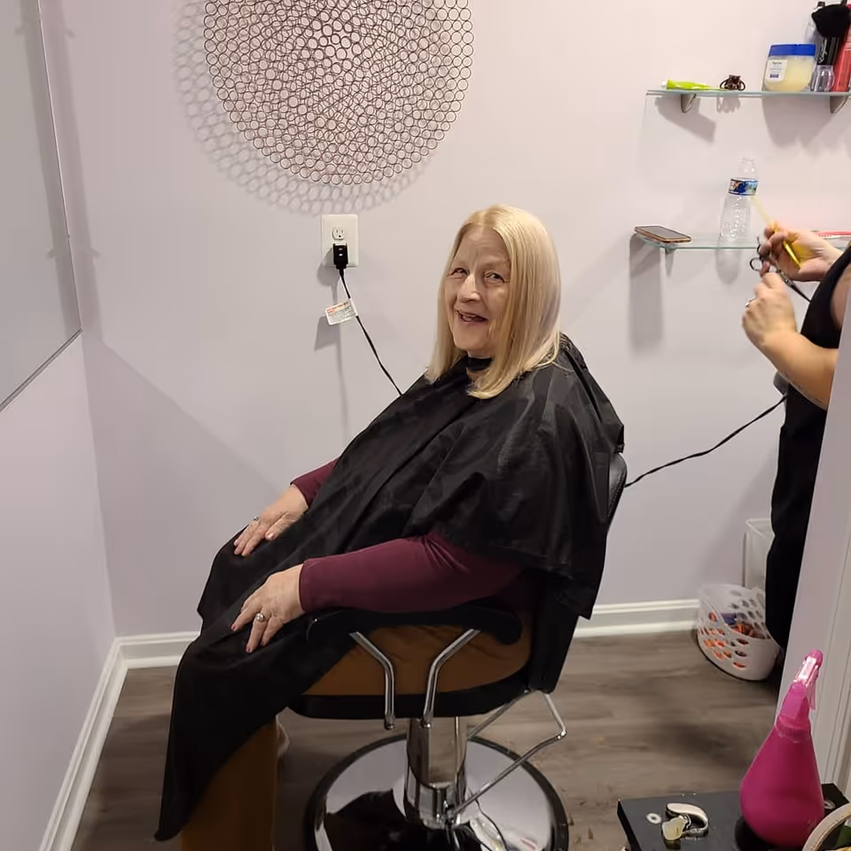 An elderly woman with blonde hair sits in a salon chair wearing a black haircutting cape. A hairstylist is standing behind her, holding scissors and a comb, preparing to cut her hair. The room has light-colored walls with a decorative circular wall piece and glass shelves holding various items including a water bottle and hair products.