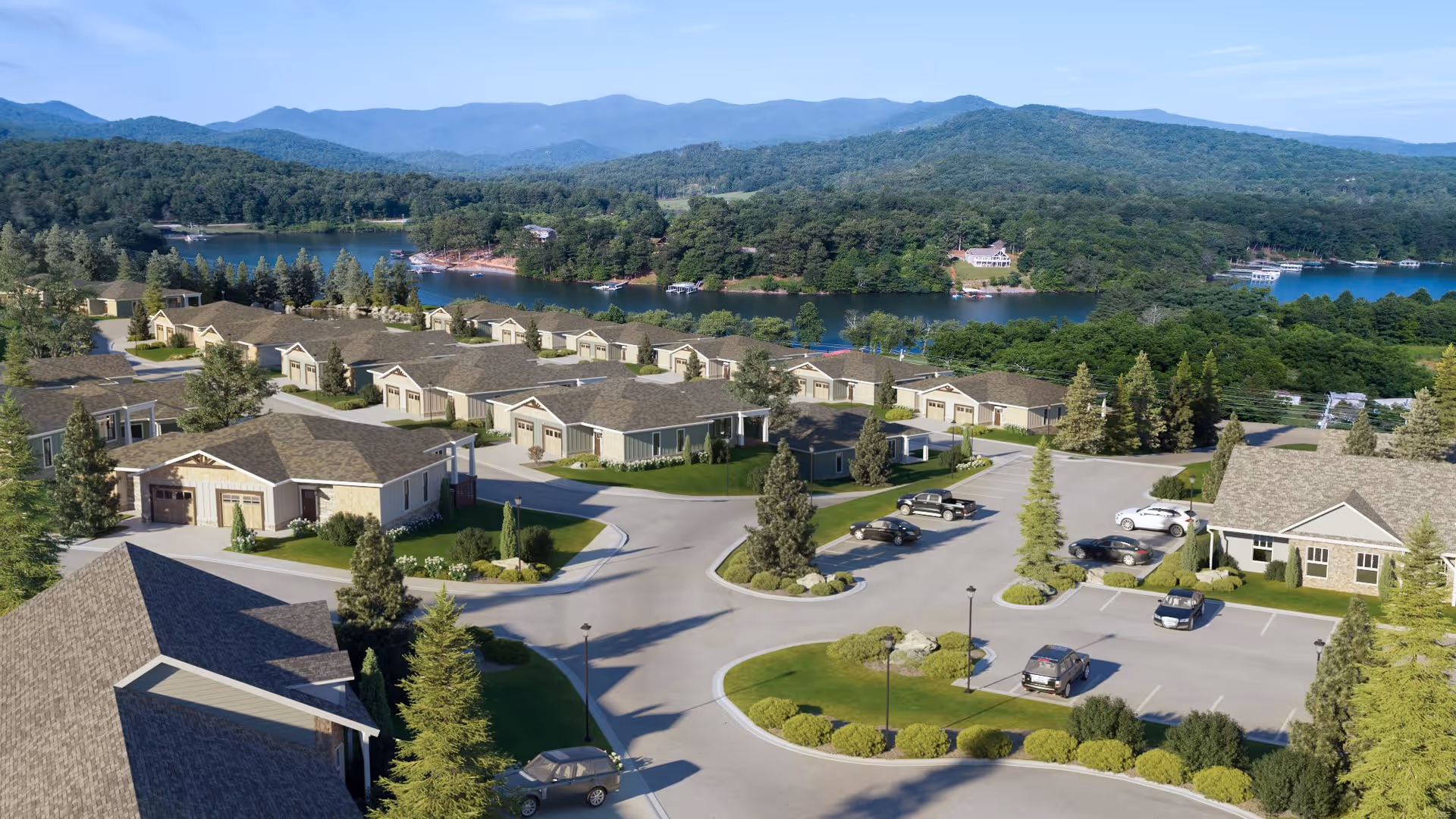 Aerial view of a residential community with single-story houses, parking areas, and landscaped greenery. The community is situated near a large body of water with forested hills and mountains in the background under a clear blue sky.