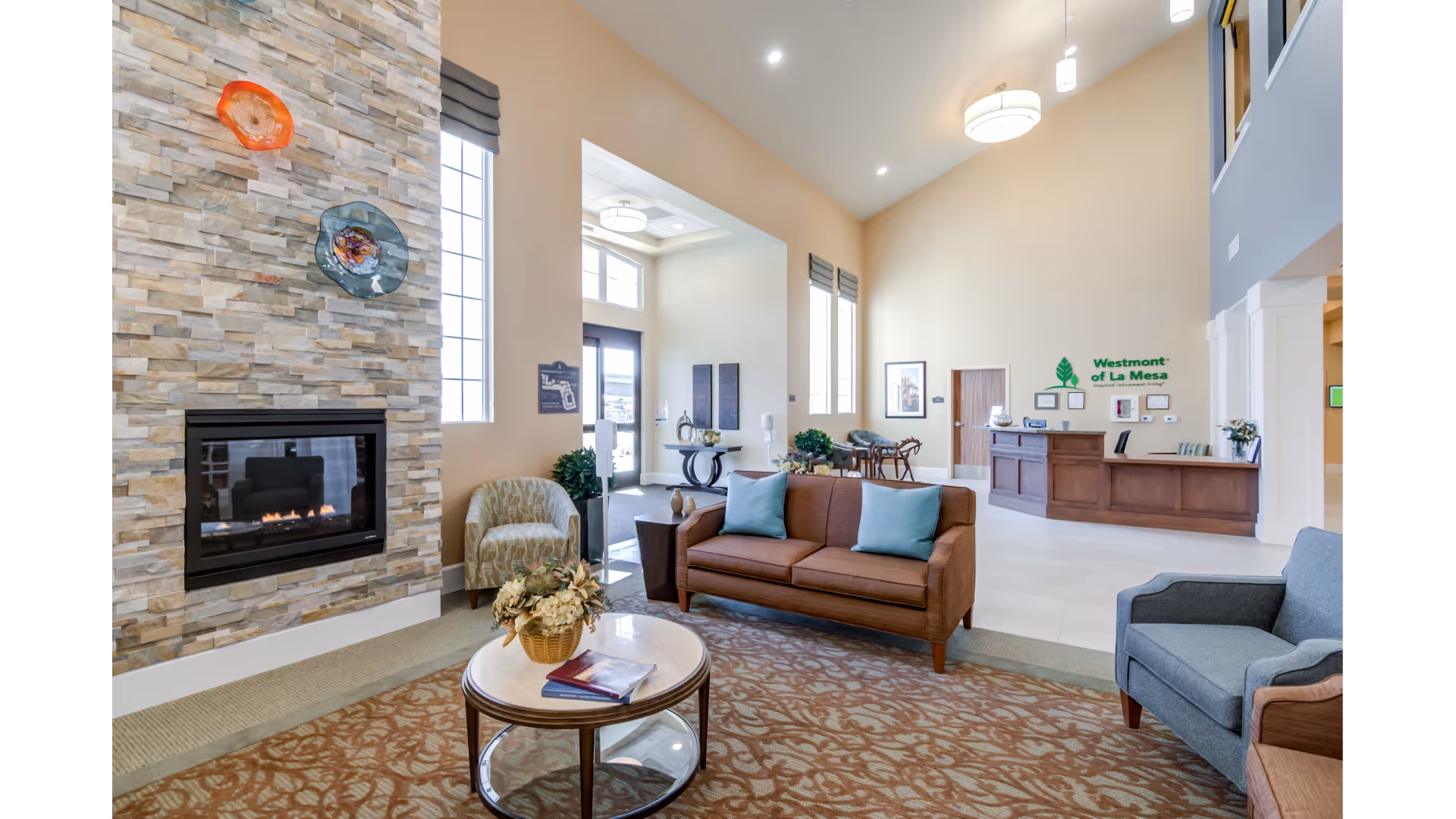 Spacious senior living lobby with seating area, fireplace, and a reception desk with signage.