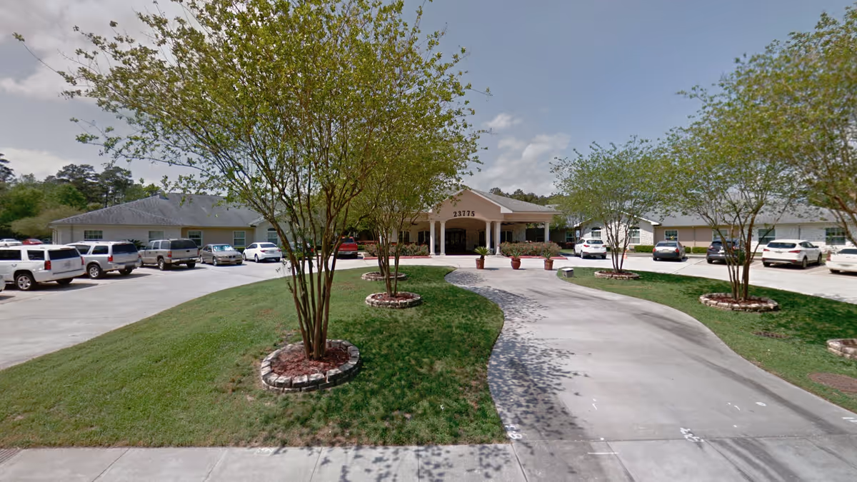 Front exterior view of Kingwood Rehabilitation & Healthcare Center showing a driveway lined with small trees and a parking area with several cars parked on both sides. The building has a covered entrance with the number 23775 displayed above it.