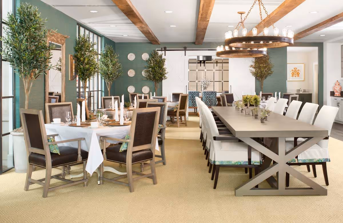 A spacious and elegant dining room with two large tables set for a meal. One table is round with a white tablecloth and dark wooden chairs with armrests, while the other is a long rectangular table with white cushioned chairs. The room features green walls, large windows, decorative plants, wooden ceiling beams, and two large circular chandeliers hanging above the tables.