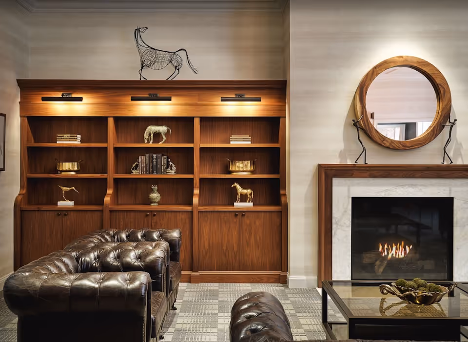 A cozy living room area featuring a dark brown tufted leather sofa and ottoman, a wooden bookshelf with decorative items including horse sculptures and books, a fireplace with a wooden and marble surround, and a round wooden-framed mirror above the fireplace.