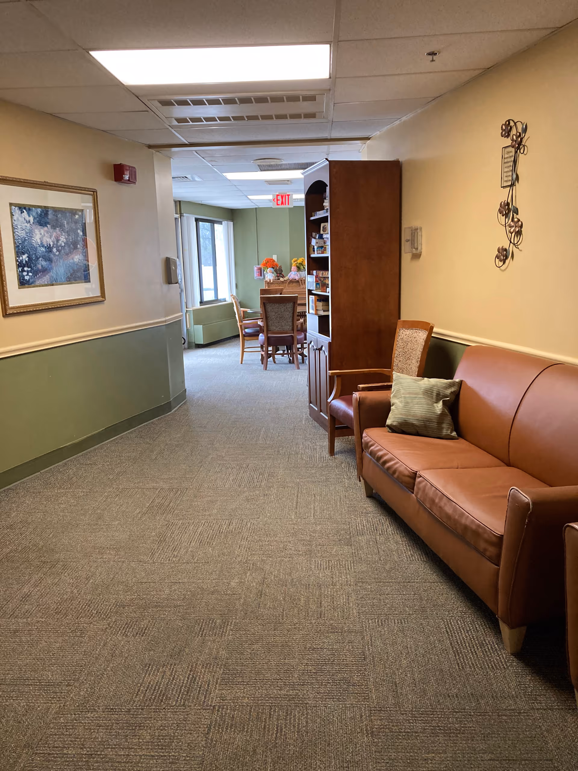 A hallway in a senior living facility with beige and green walls, carpeted floor, and fluorescent ceiling lights. On the right side, there is a brown leather couch with a green pillow and a wooden chair next to it. Further down the hallway, there is a wooden bookshelf and a dining area with a table and chairs near windows letting in natural light. A framed floral painting hangs on the left wall.