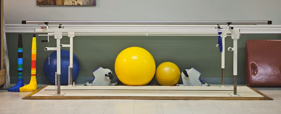 Physical therapy equipment including parallel bars for walking exercises, two exercise bikes, two large yellow exercise balls, a blue exercise ball, colorful foam foot models, and a brown padded mat against a green wall.
