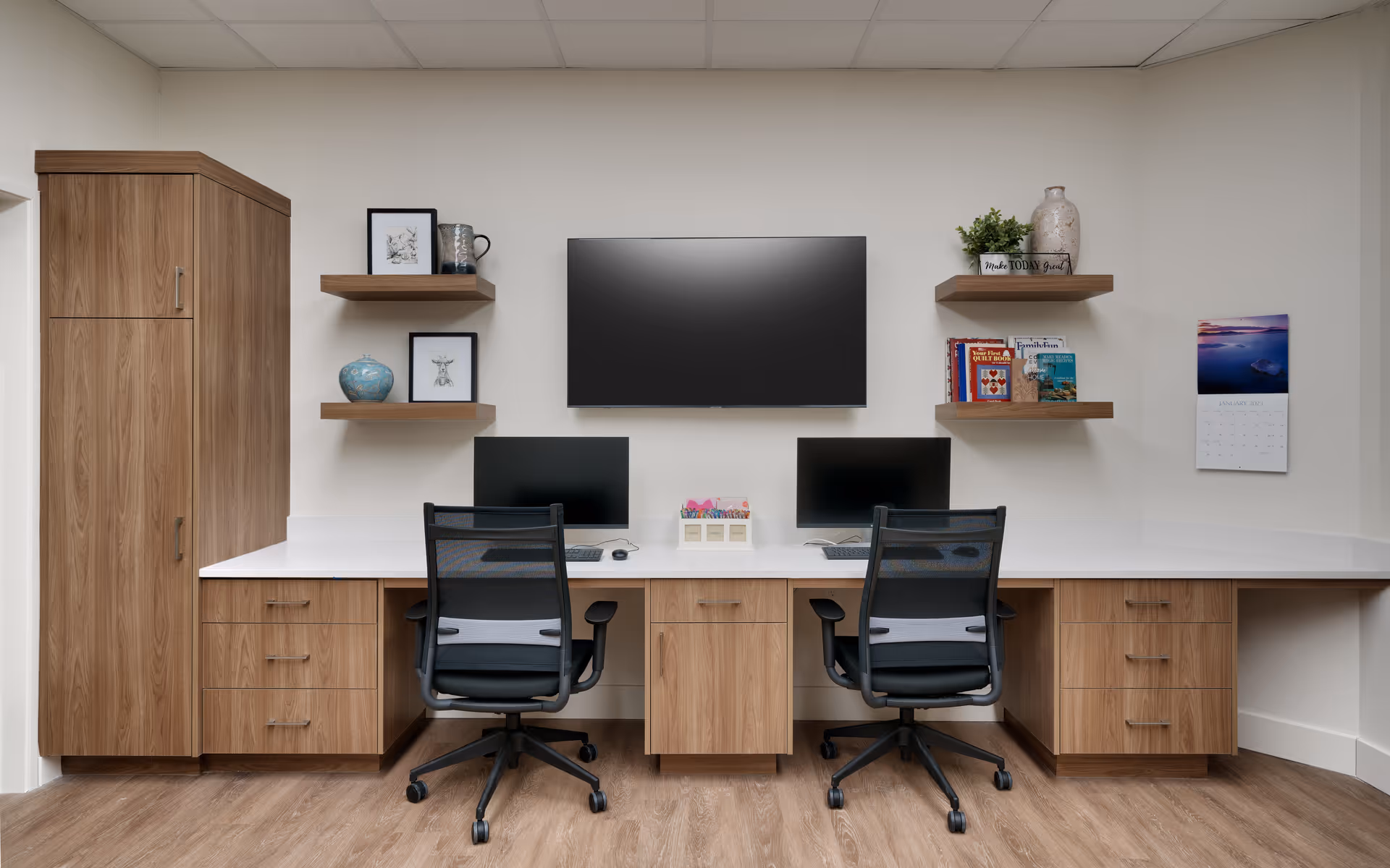 A modern office workspace with two black mesh office chairs in front of a long white desk with wooden cabinets and drawers. Two computer monitors are placed on the desk, with a large flat-screen TV mounted on the wall above. Wooden floating shelves on either side of the TV hold decorative items, books, and plants. A calendar is hanging on the right wall.