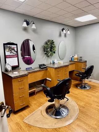 Interior view of a salon area with two black salon chairs in front of wooden counters with mirrors mounted on the wall. The wall is painted gray and decorated with a green wreath. There are light fixtures above each mirror and various salon supplies on the counters.