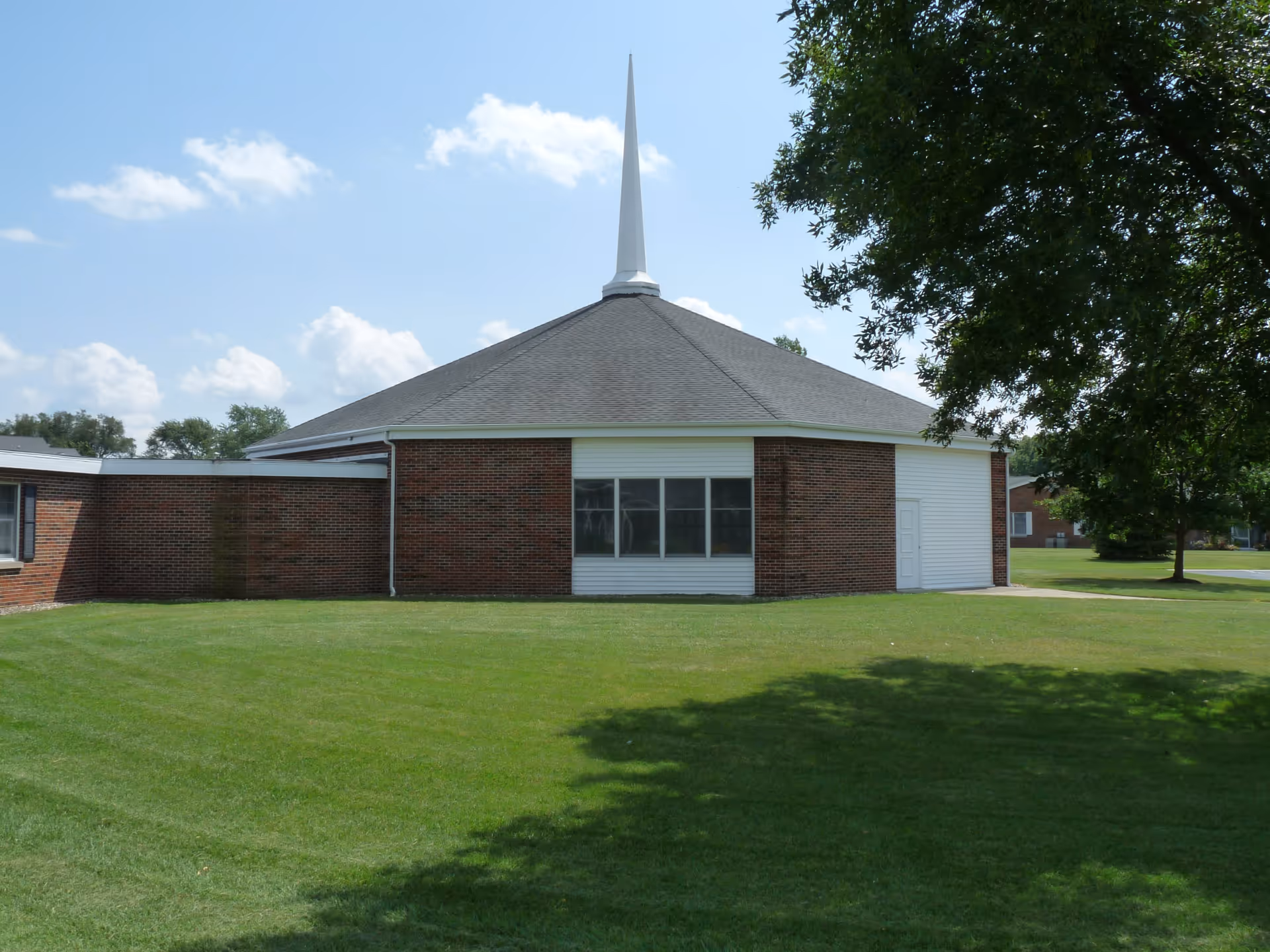 Low brick building with a conical roof topped by a white spire, set on a green lawn with trees under a blue sky.