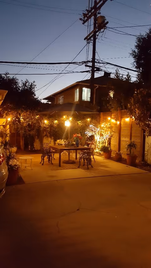 Outdoor patio area at dusk with string lights hanging above a table and chairs. There are potted plants and a wooden fence surrounding the space, with a house and power lines visible in the background.