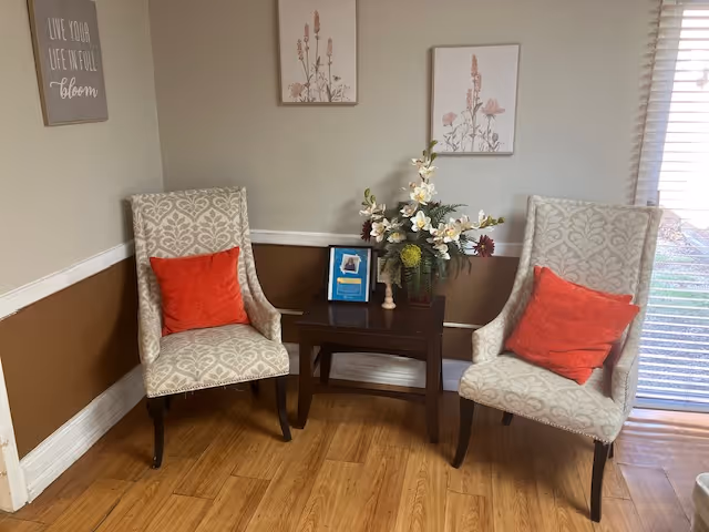 Two patterned armchairs with red pillows flank a small wooden table holding a floral arrangement and a framed picture in a cozy seating nook.